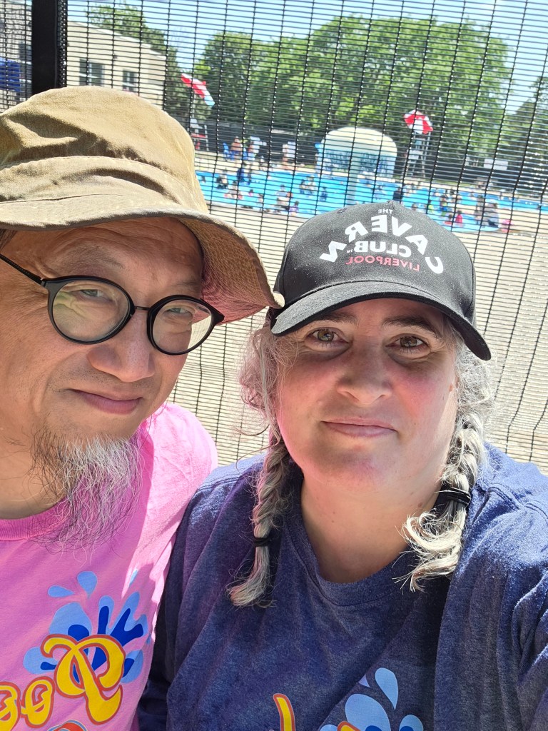 A selfie of a man and woman in front of a swimming pool, wearing colorful shirts and hats. The pool area is visible in the background with sunny weather and people enjoying the water.