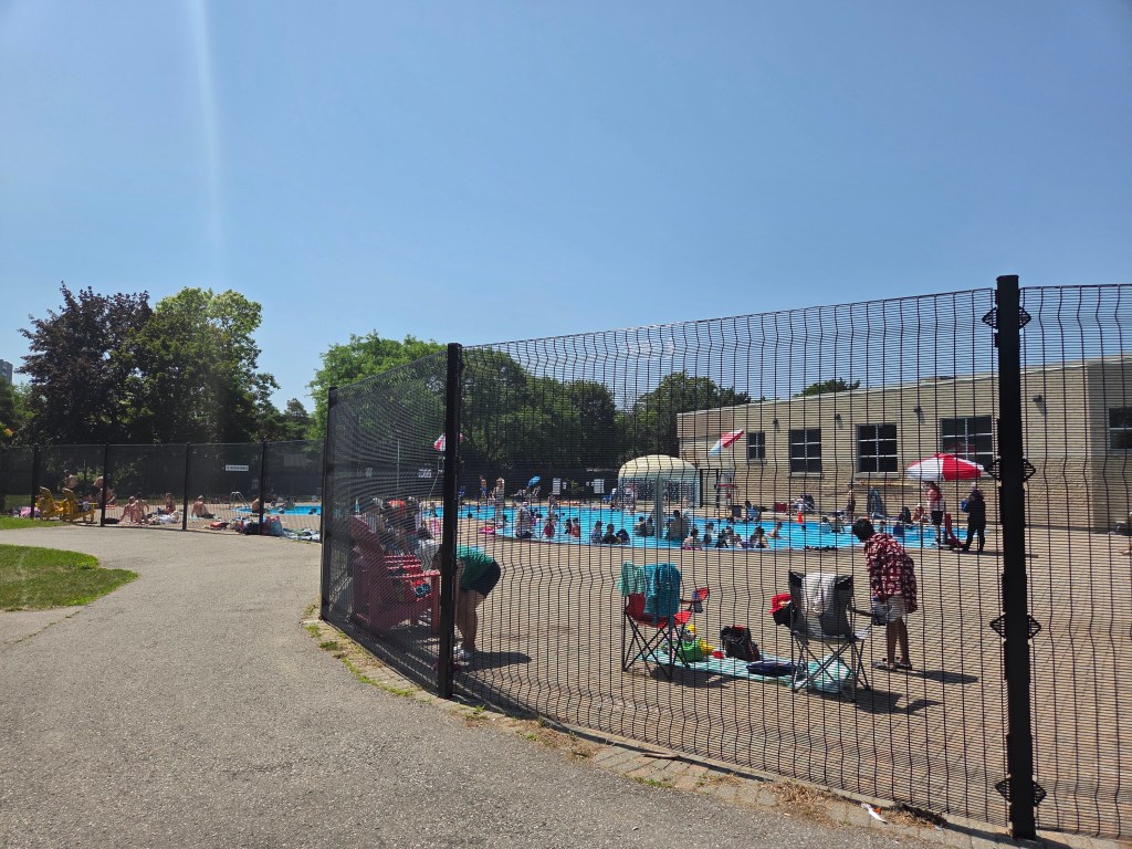 A community swimming pool surrounded by a black fence, filled with people enjoying a sunny day. Several colorful umbrellas and lawn chairs are set up around the pool area.