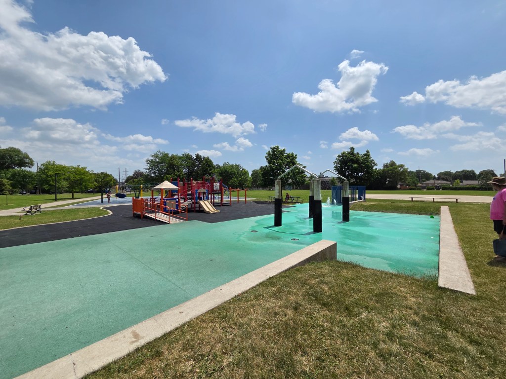A playground during the day featuring a colorful play structure with slides, surrounded by green grass and trees. A water play area with several spray features is visible in the foreground, with clear blue skies and fluffy clouds in the background.