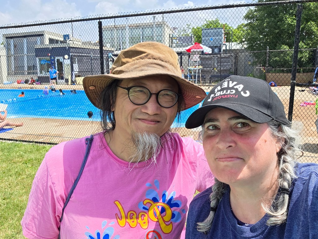 A man and a woman smiling for a selfie at a swimming pool, with people swimming in the background and a sunny sky.