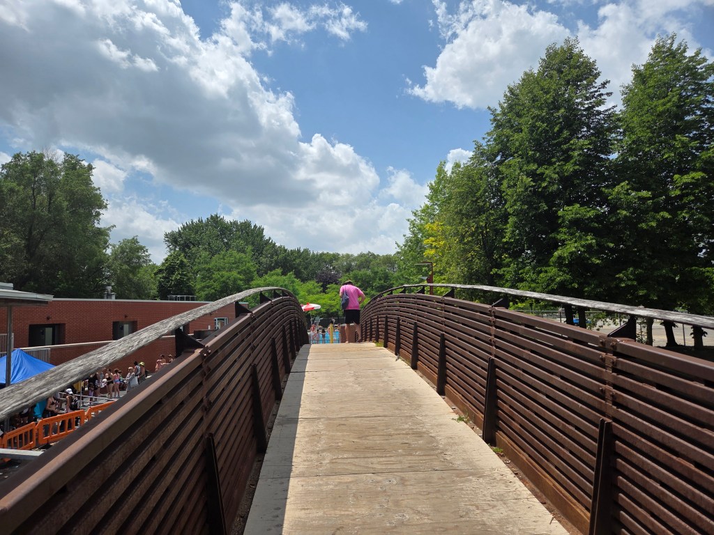 View of a wooden pedestrian bridge with a person standing at the end, surrounded by trees and blue skies with fluffy clouds.