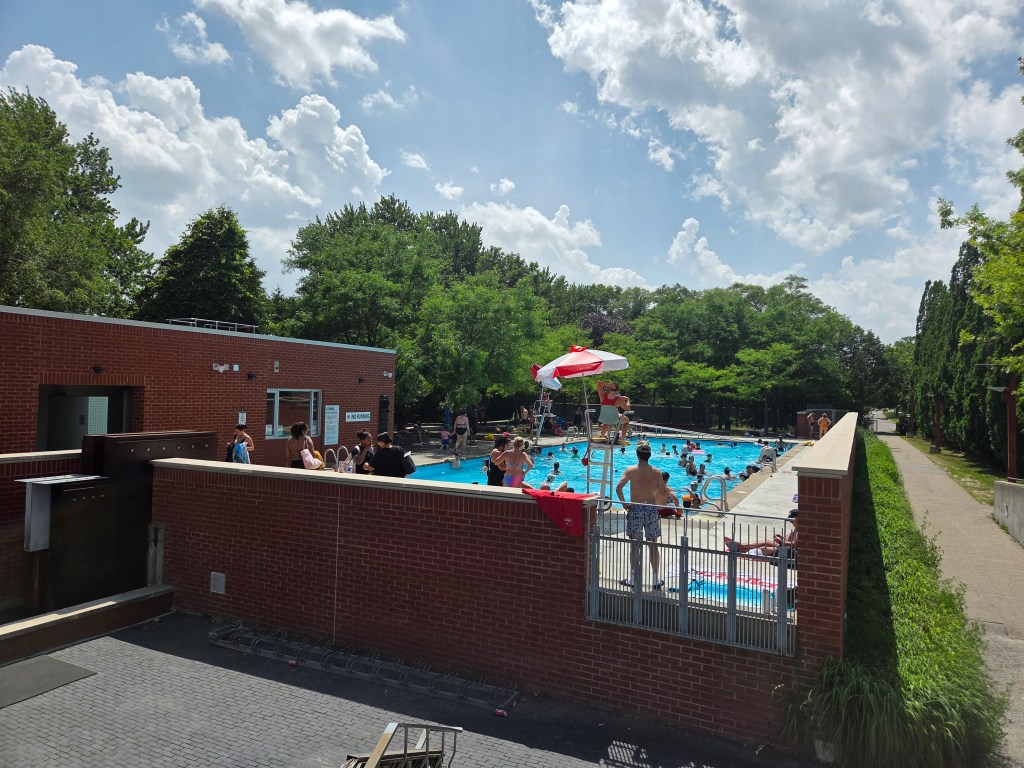 An outdoor swimming pool scene with many people enjoying the water on a sunny day, surrounded by green trees and a brick building nearby.