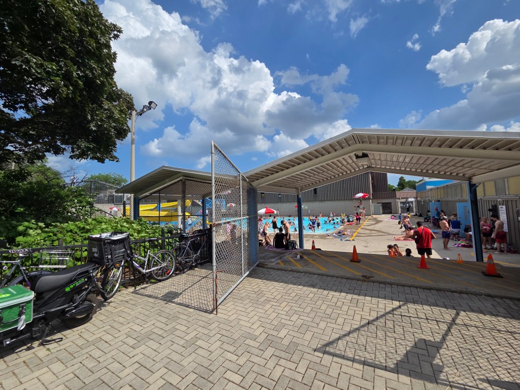 A view of a public swimming pool area filled with people enjoying the sun and water, with large umbrellas providing shade, surrounded by greenery and bicycles parked near a chain-link fence.