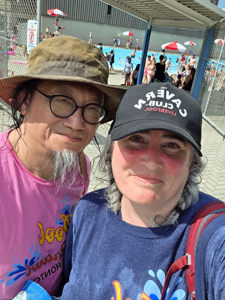 A man and a woman posing for a selfie at a public pool on a sunny day. The man wears a camouflage hat and a pink shirt, while the woman sports a black cap and a dark blue shirt. In the background, people are enjoying the pool with umbrellas and swimming.