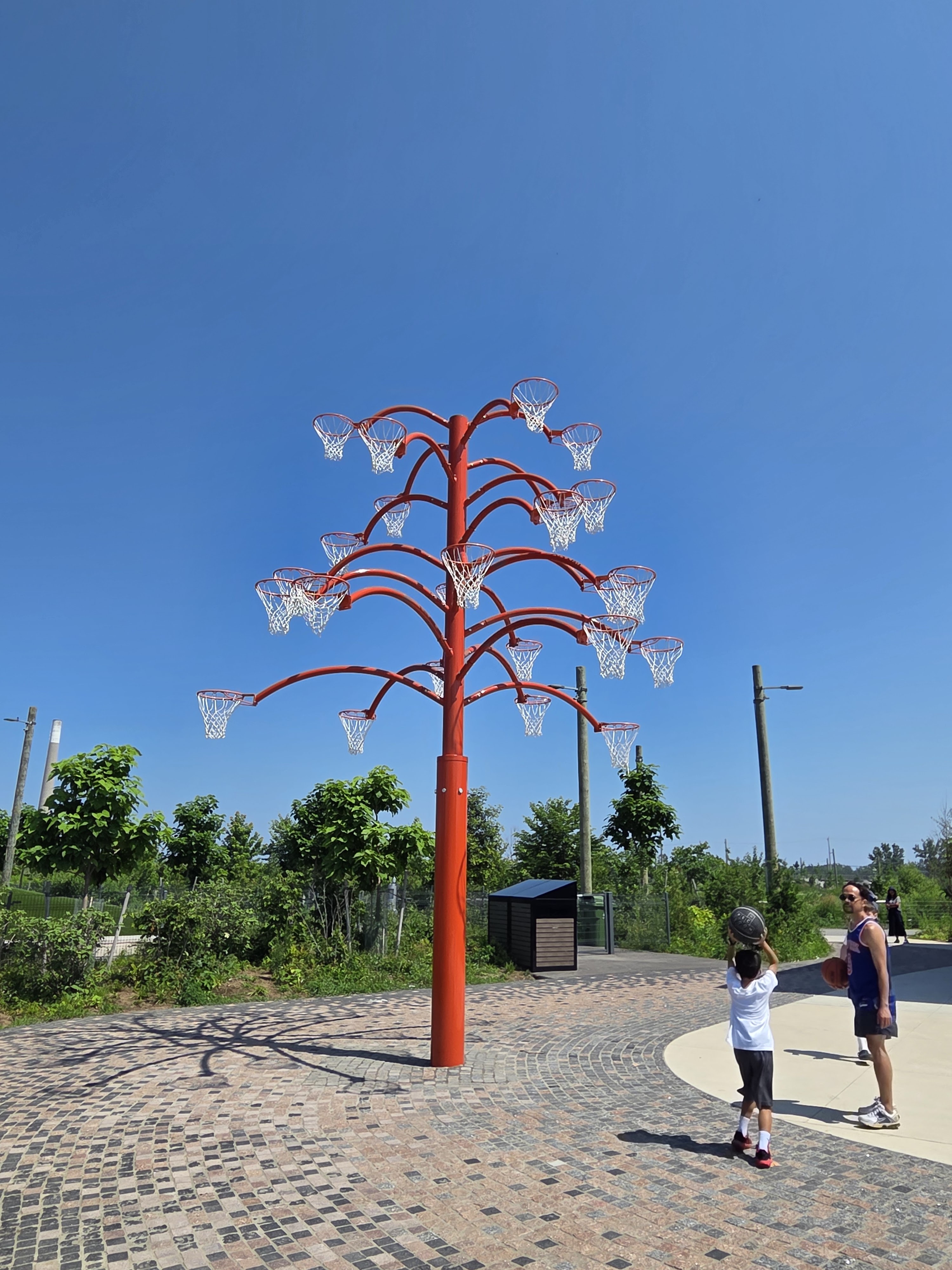 An innovative basketball hoop tree structure with multiple hoops, set against a clear blue sky, with two people playing basketball nearby.