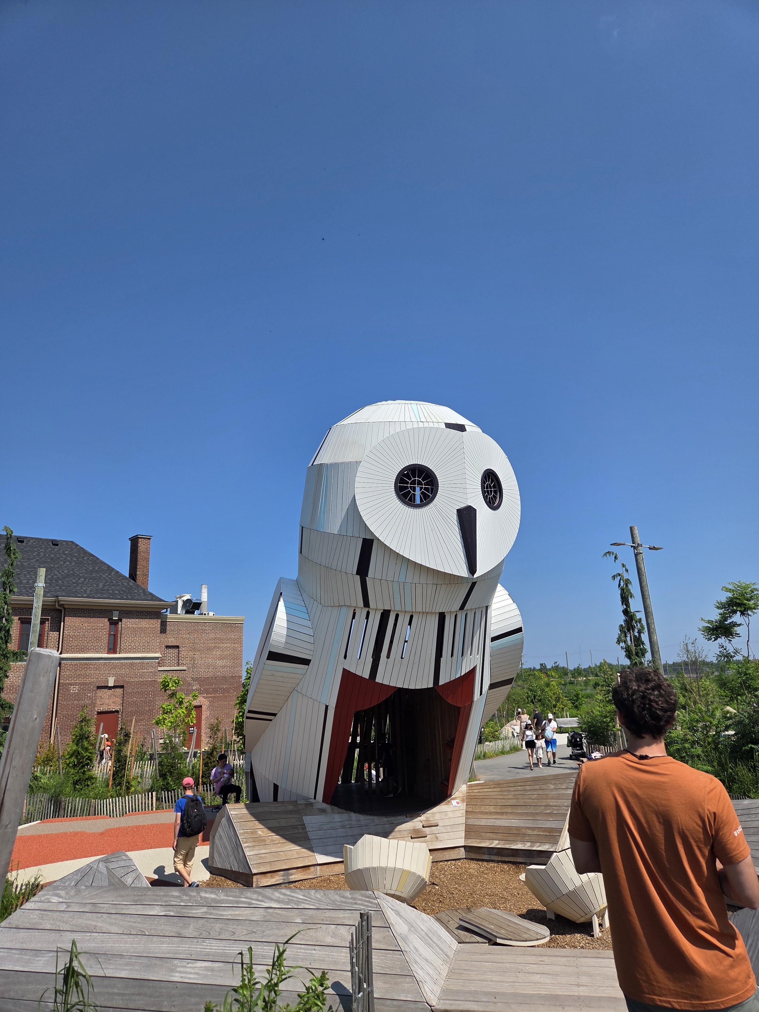 A large, whimsical owl-shaped structure with a white exterior, set against a clear blue sky. People are walking nearby, and there are wooden pathways and landscaping around the installation.
