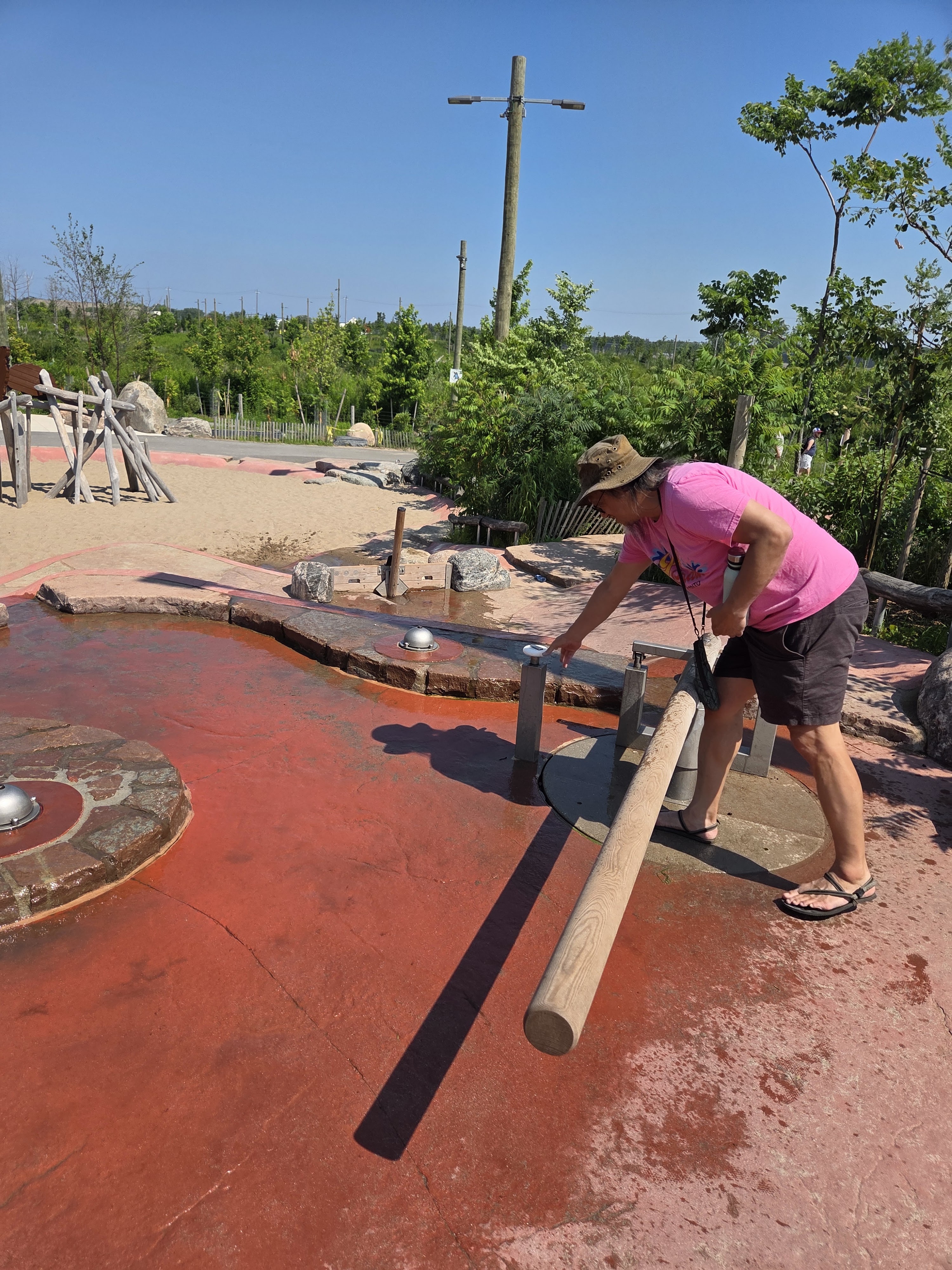 A person interacting with a water play feature at a park, manipulating a large wooden pole near a circular water basin. The surroundings include greenery, sandy areas, and a clear blue sky.