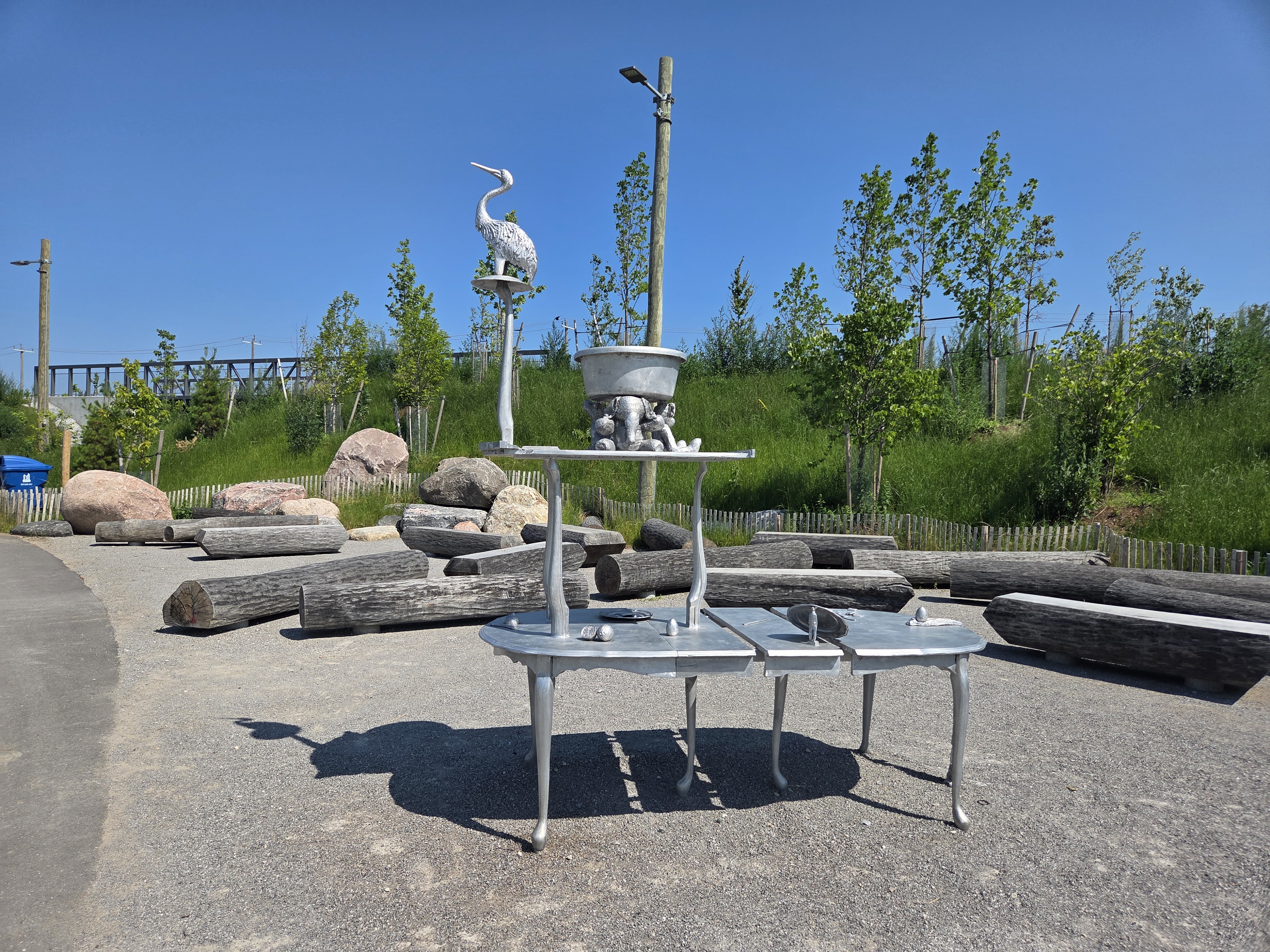 A whimsical outdoor sculpture featuring a table with silver-colored legs, a bucket, and a crane figure perched on top, set in a green park surrounded by rocks and logs.