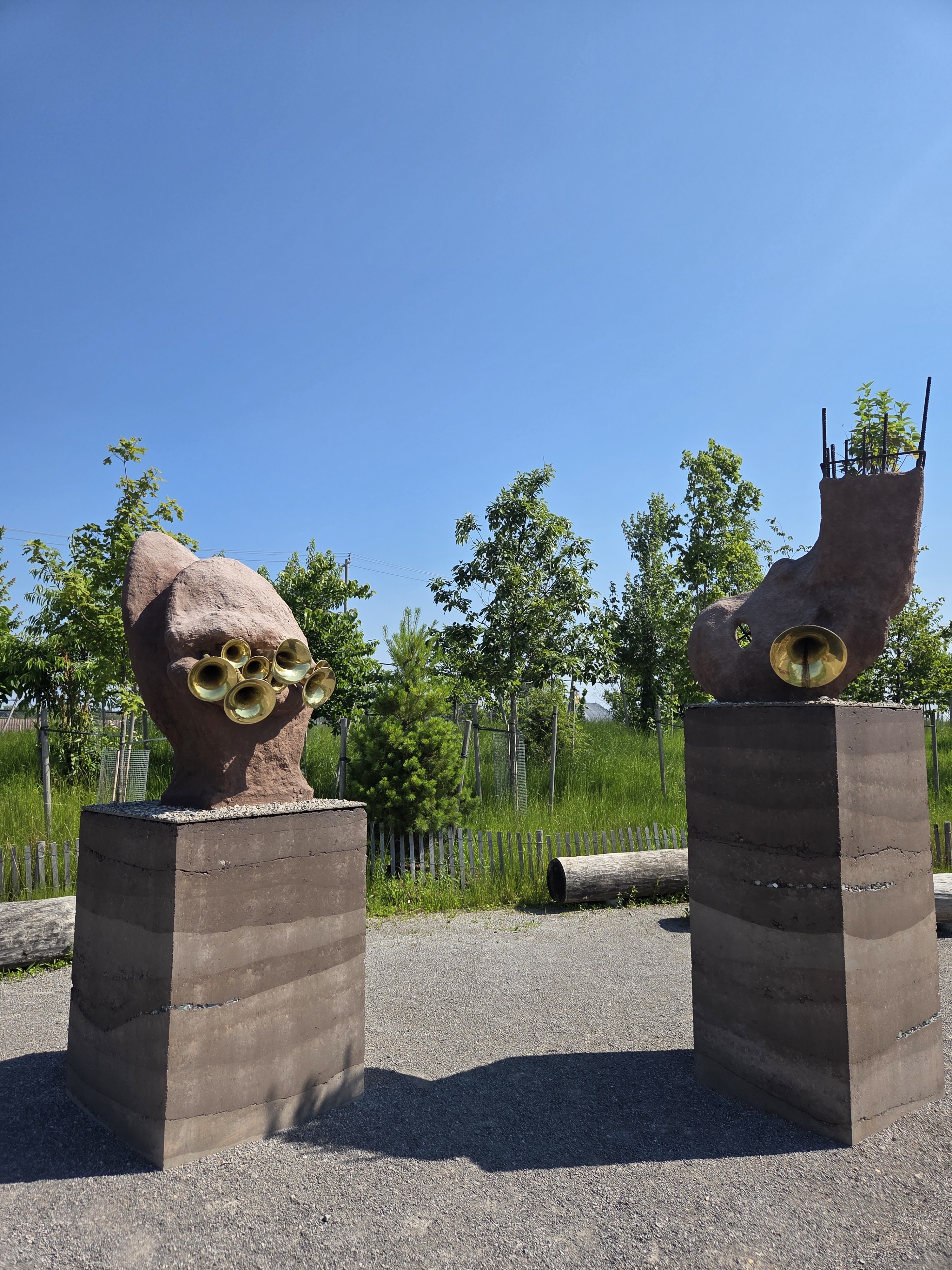 Two abstract sculptures made of stone and featuring horn-like elements, set against a clear blue sky and surrounded by greenery.
