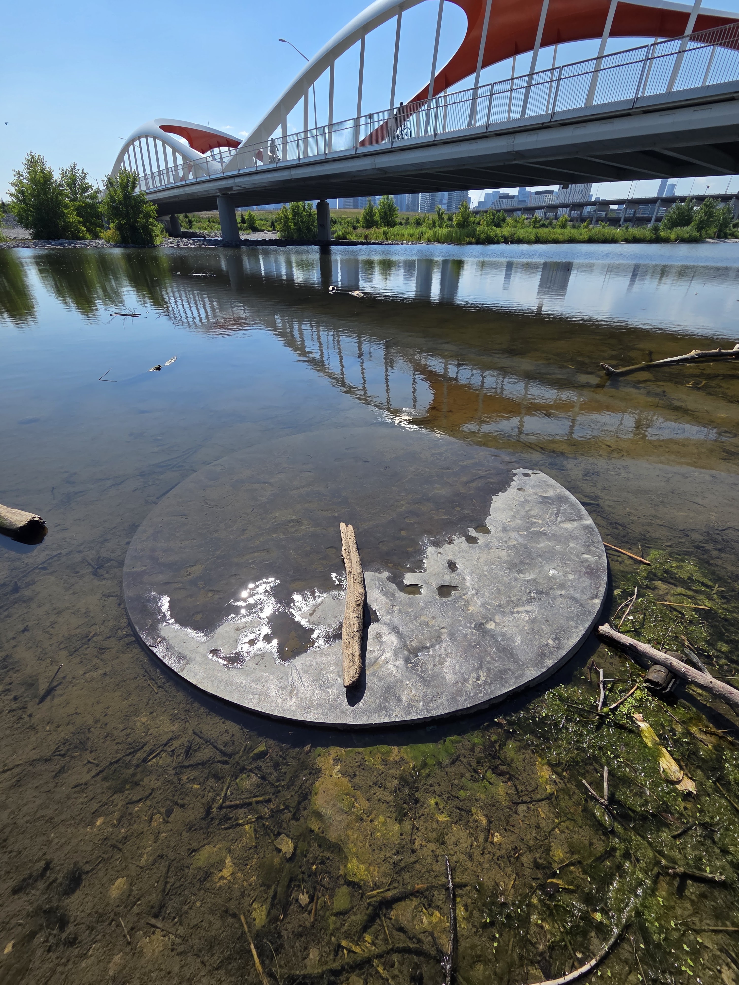 A circular platform in a river with a stick resting on it, surrounded by clear water and reflecting the blue sky, with a modern bridge in the background.