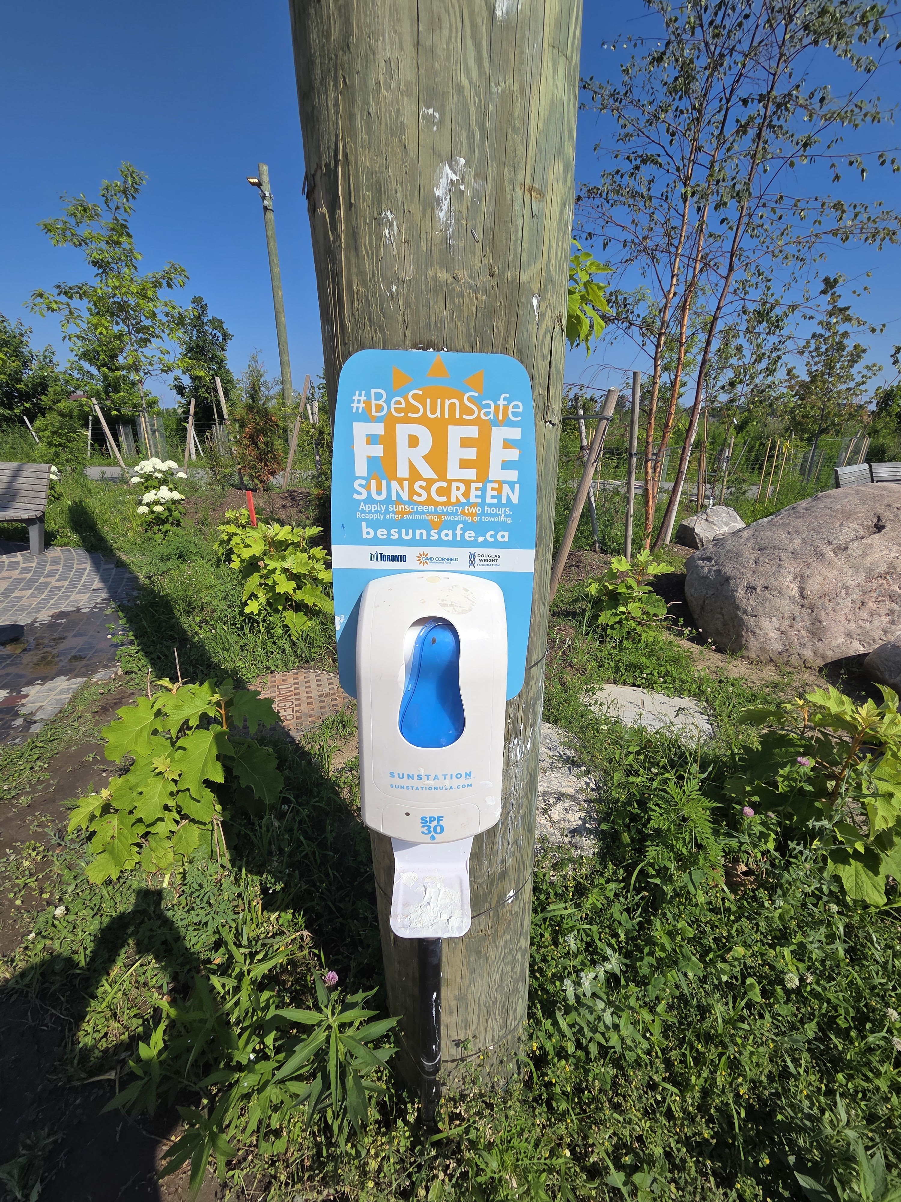 A post-mounted sunscreen dispenser labeled 'FREE SUNSCREEN' with 'BeSunSafe' campaign message, surrounded by greenery and blue sky.