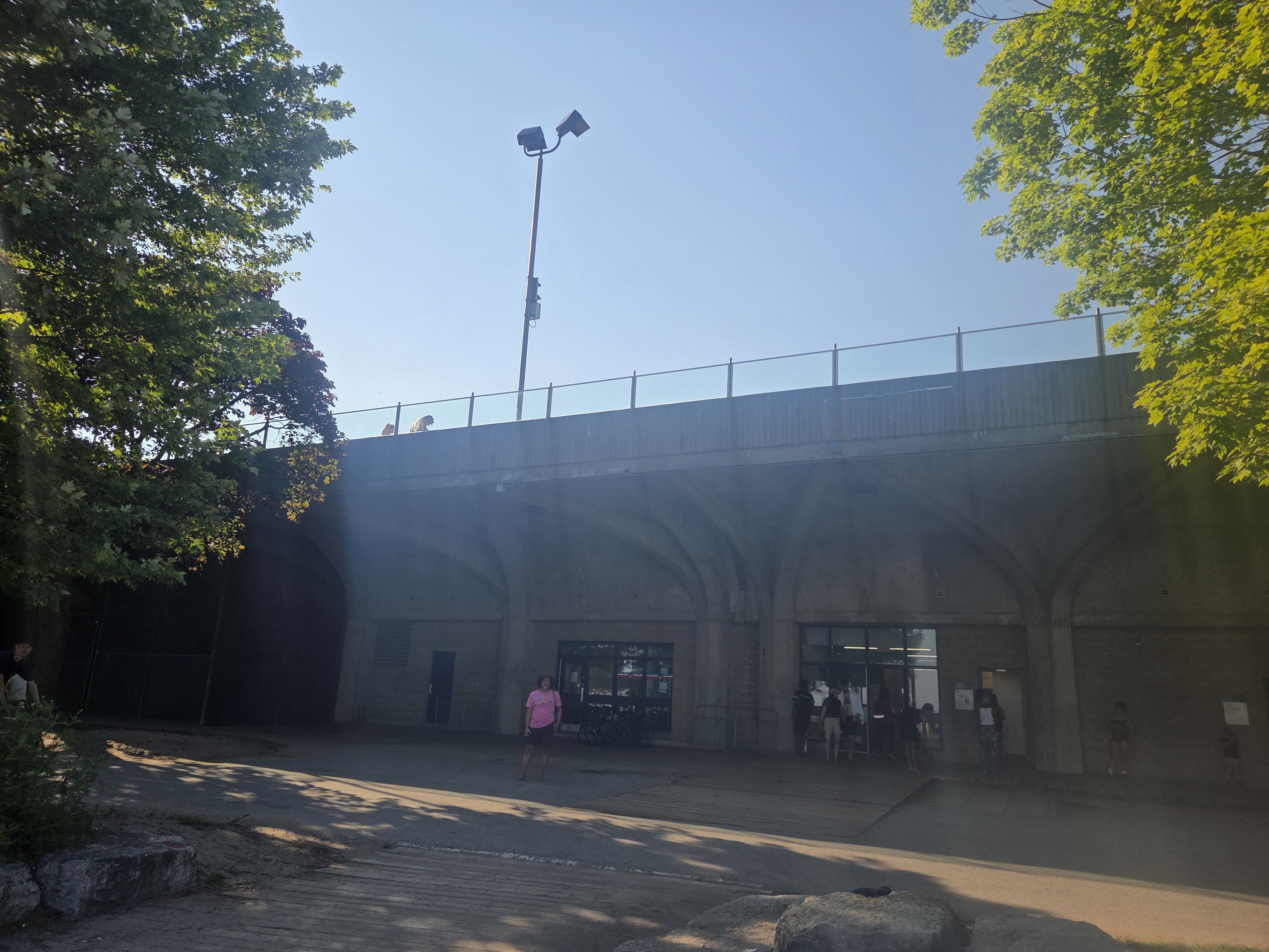 A concrete structure with an arched design, featuring a glass entrance and visible people gathering outside. There are trees on the left side and a clear blue sky above.