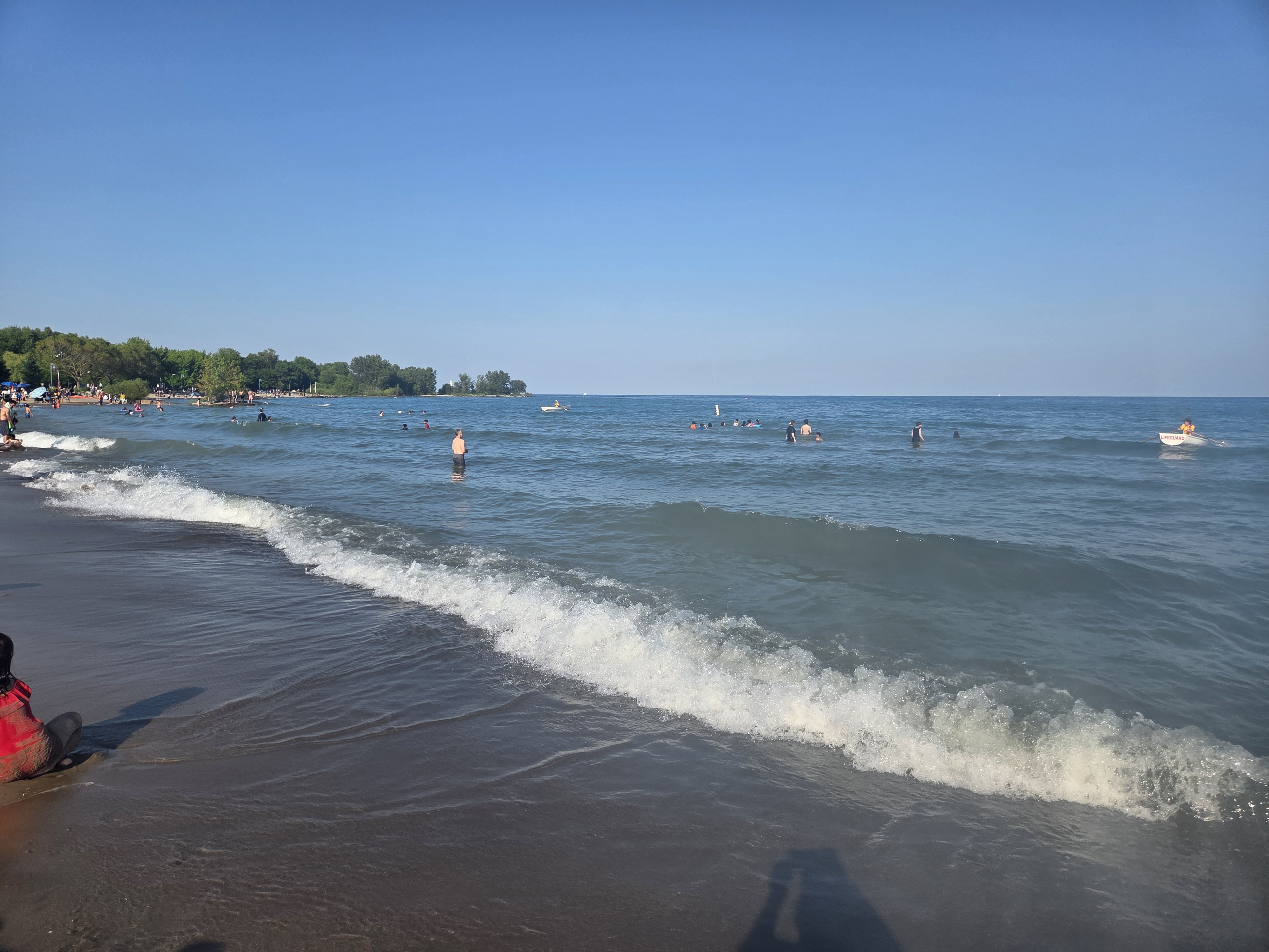 A beach scene with people swimming and enjoying the water, gentle waves lapping at the shoreline under a clear blue sky.