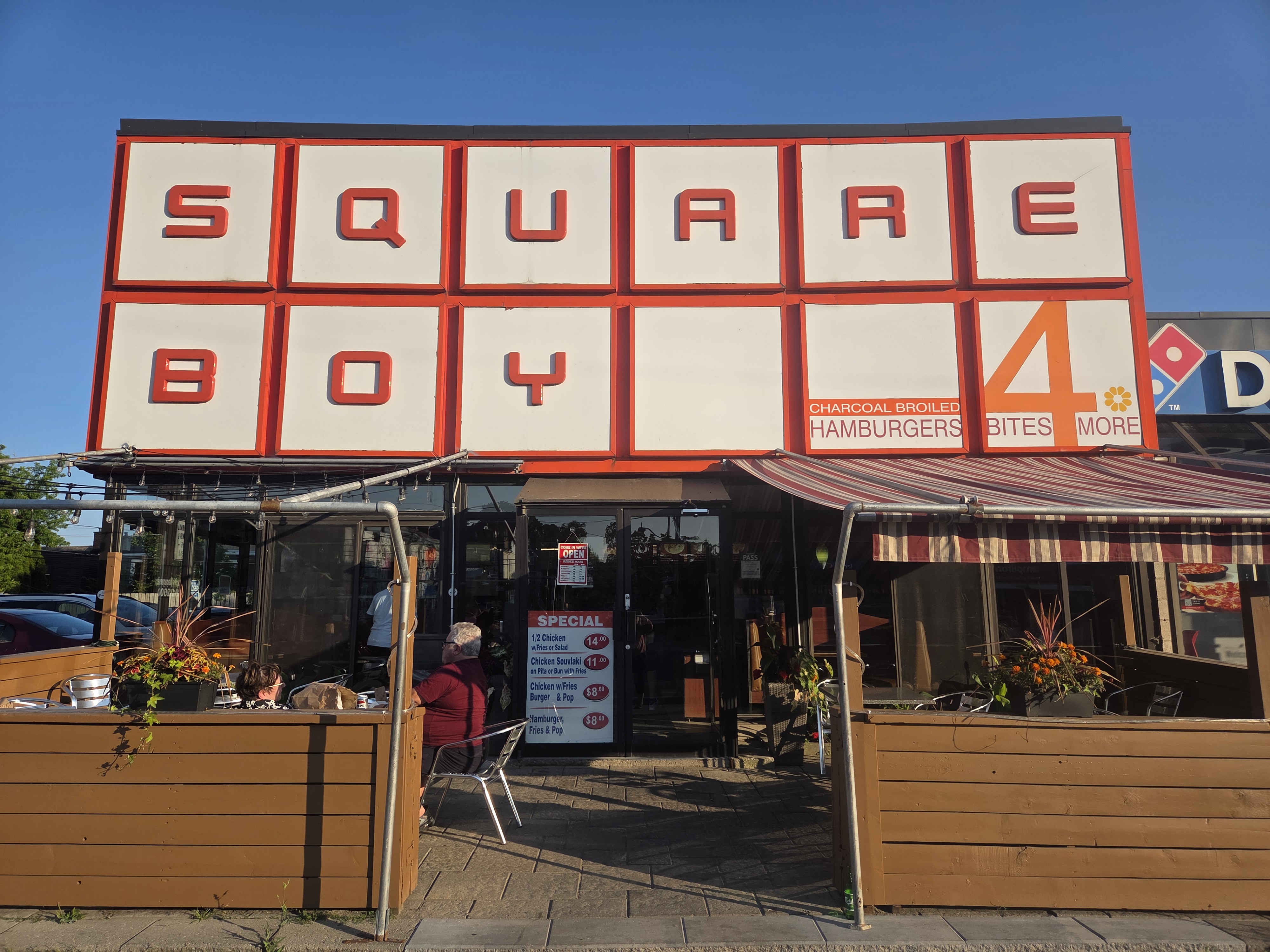 Front view of Square Boy restaurant featuring bold lettering and a sign advertising charcoal broiled hamburgers, with outdoor seating visible.