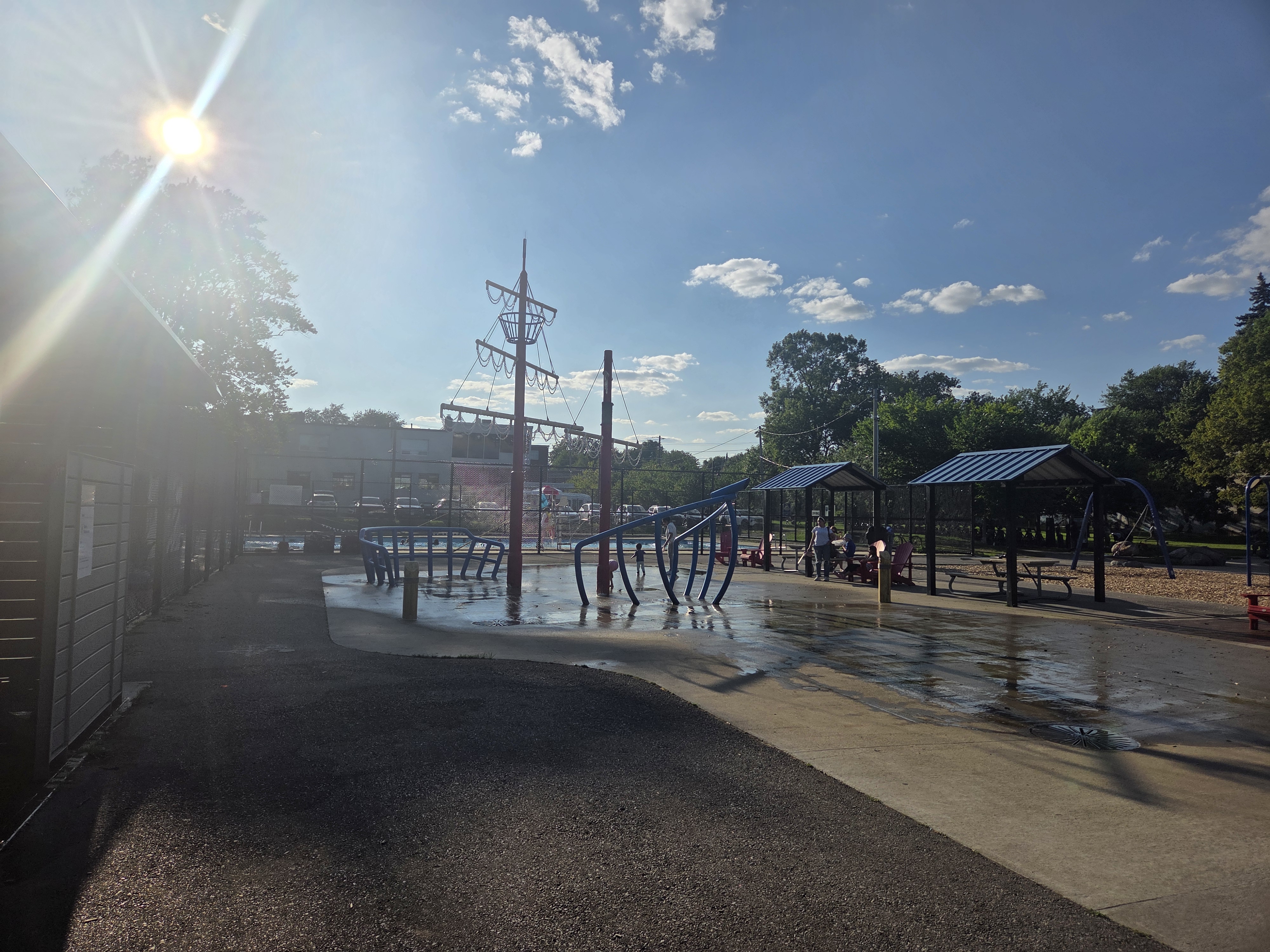 A playground scene featuring blue climbing structures, water features on a concrete surface, picnic shelters, and children playing in the background under a sunny sky.