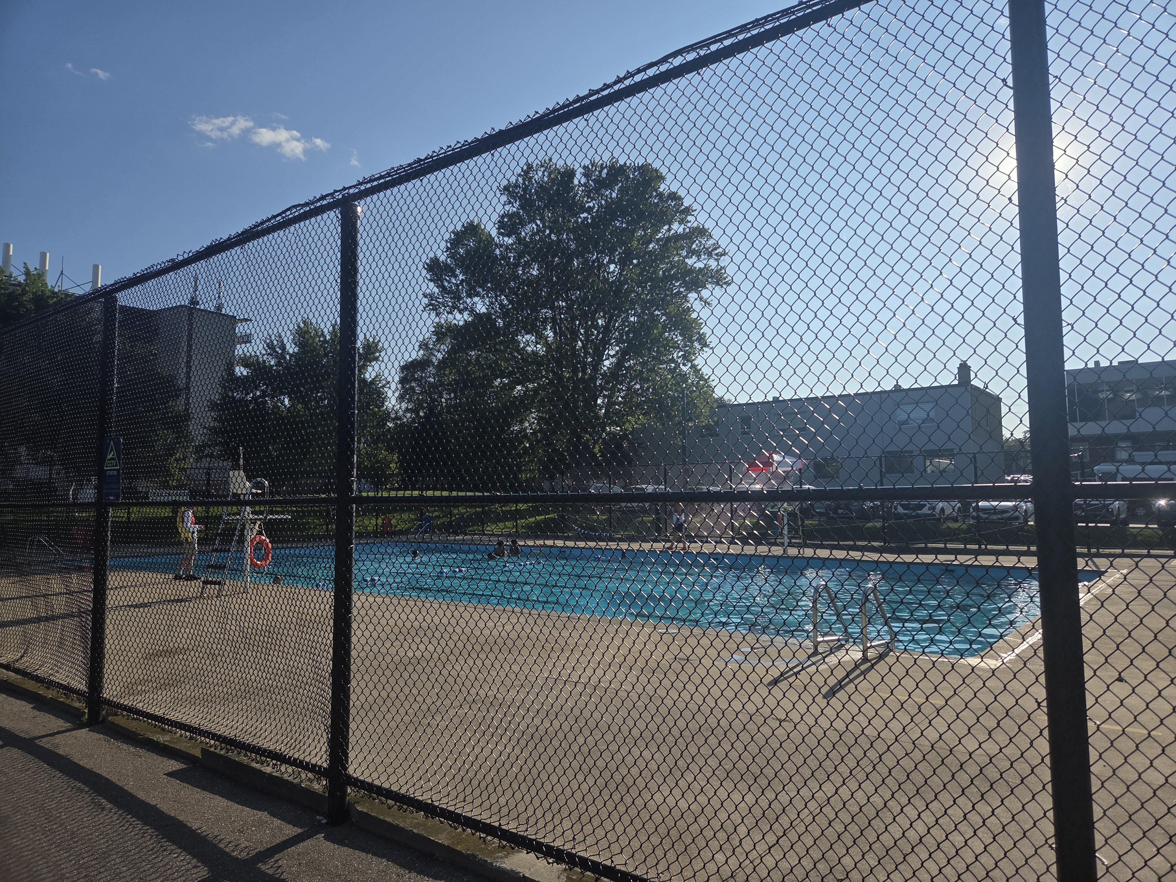 A view of a swimming pool enclosed by a chain-link fence, with a lifeguard station visible and people swimming on a sunny day.