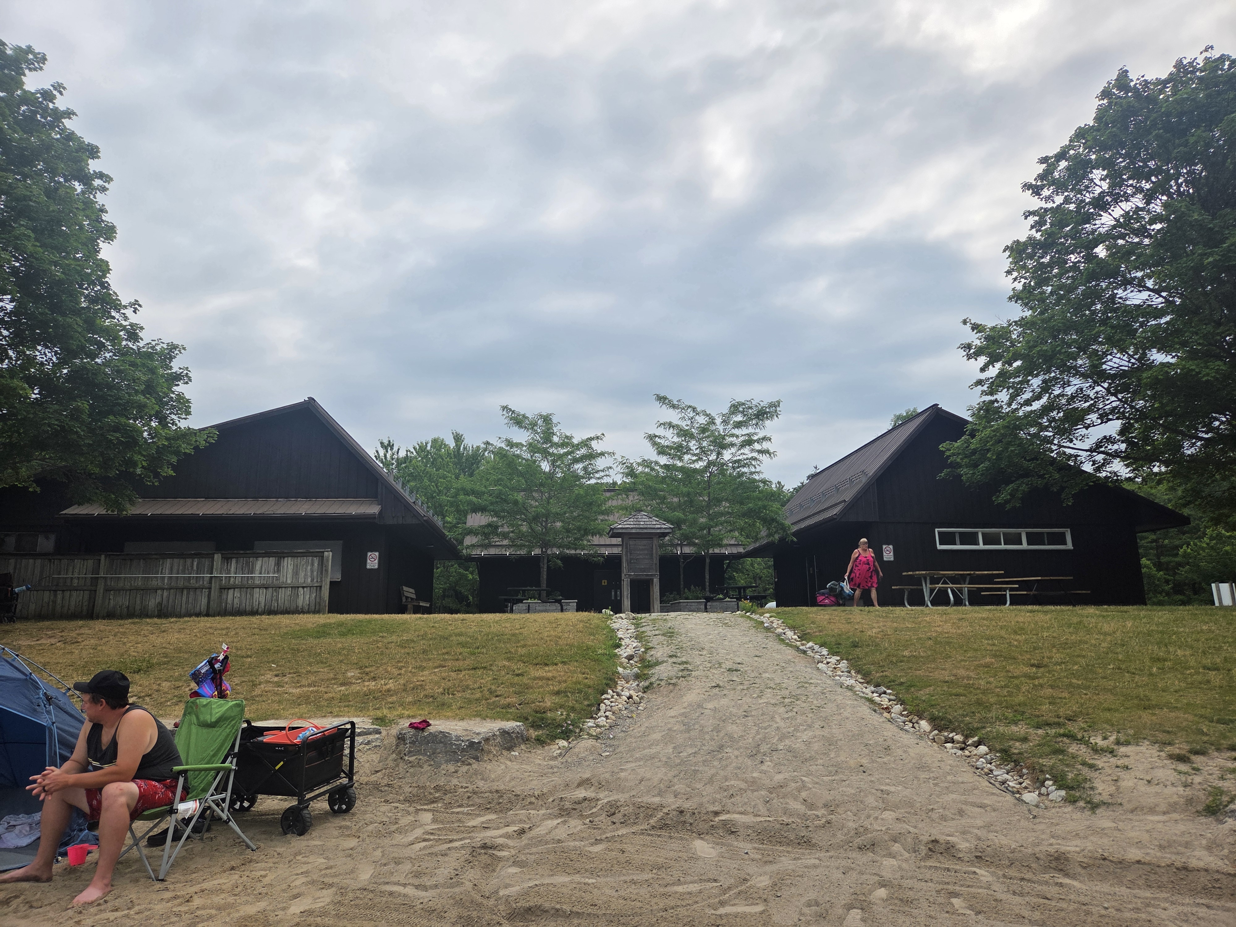 A view of a black wooden building complex with a sandy path leading to it, surrounded by green trees and cloudy skies. A person sits in a chair on the left, while another person in a pink dress walks towards the building.