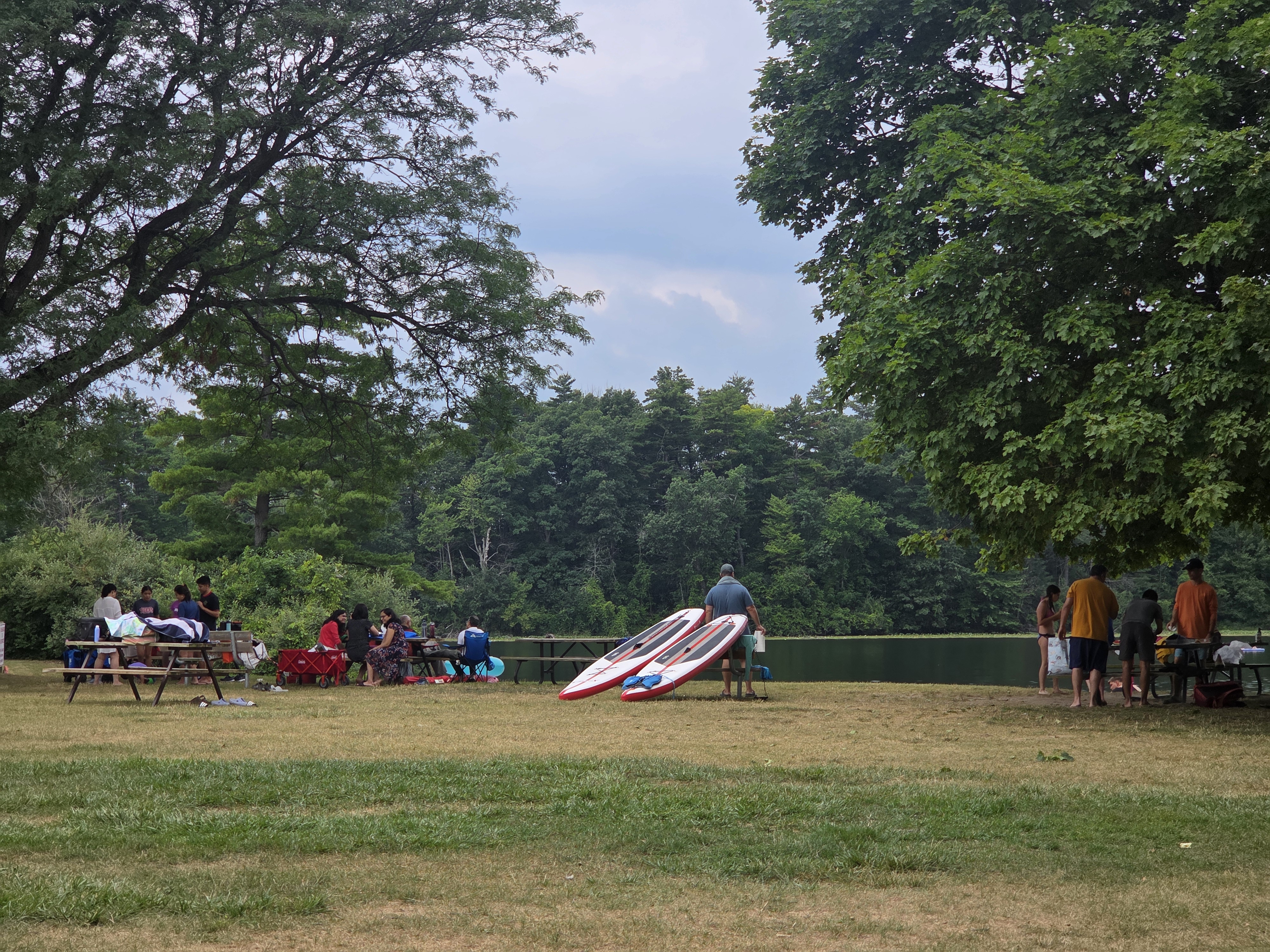 A group of people gathered by a lake with picnic tables and paddleboards, enjoying a day outdoors in a park setting.