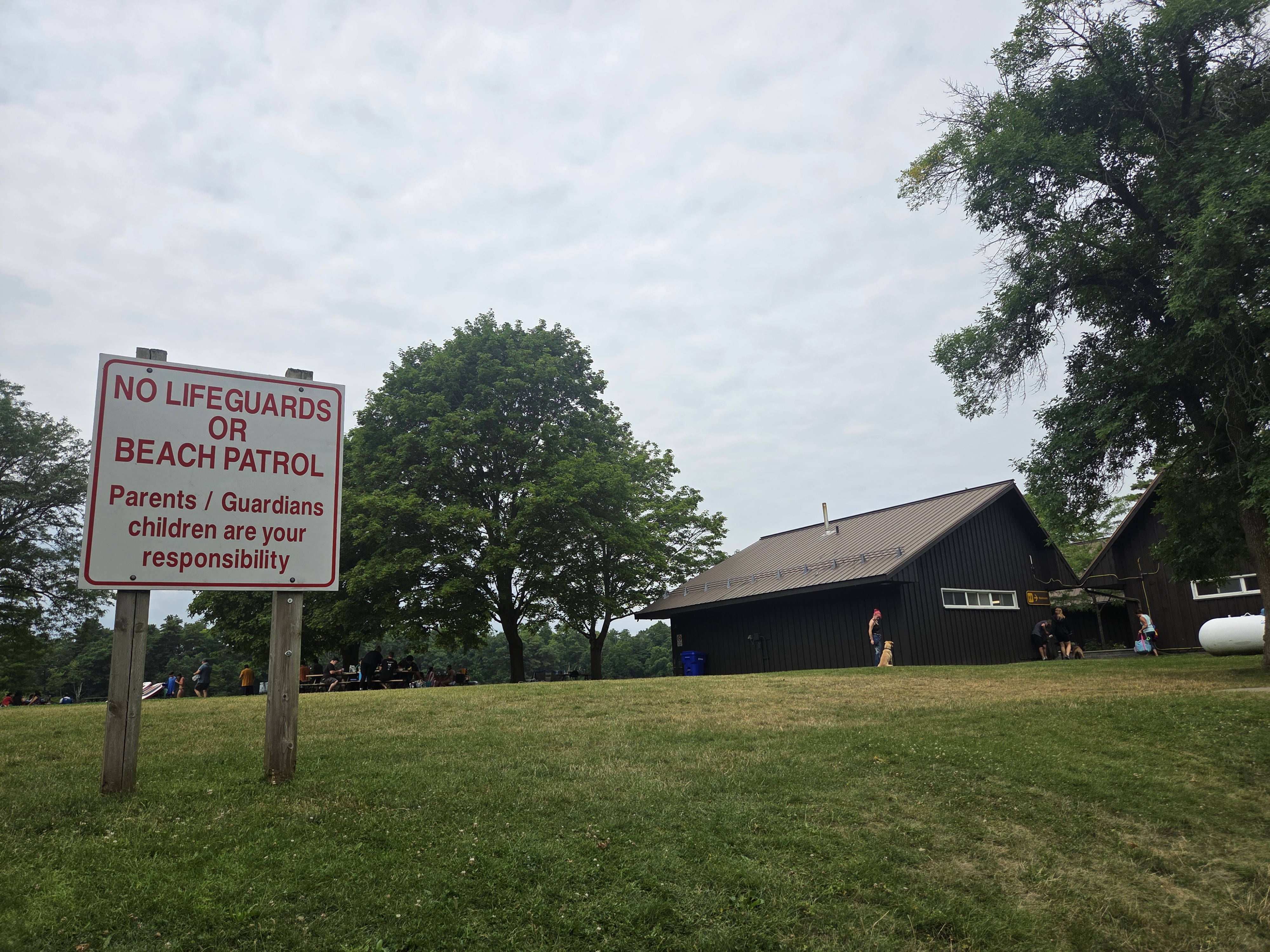 Sign indicating 'No Lifeguards or Beach Patrol' with a statement about parental responsibility for children, with green trees and a building in the background.
