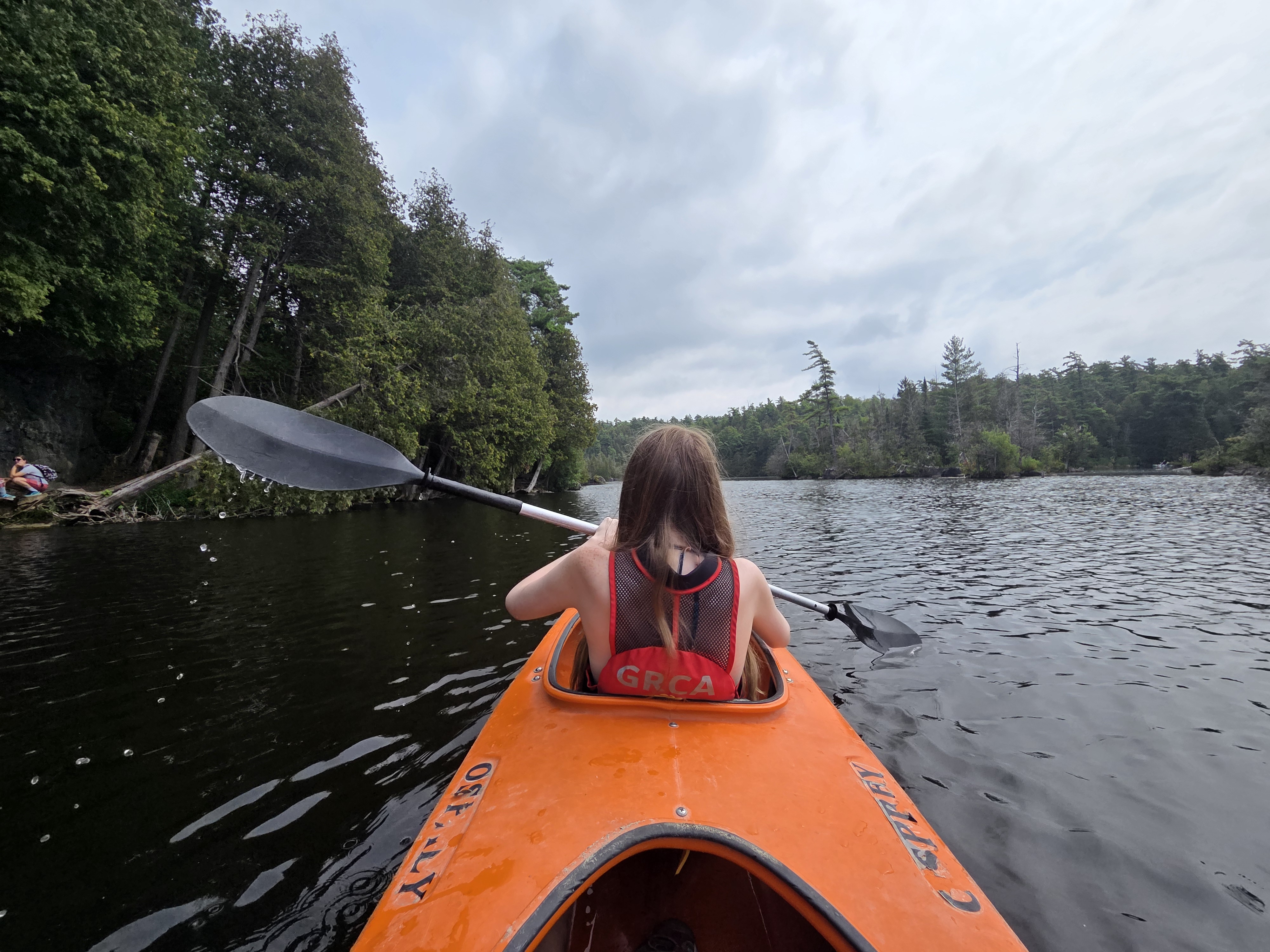 A person kayaking on a calm river surrounded by green trees and a cloudy sky, seen from behind.