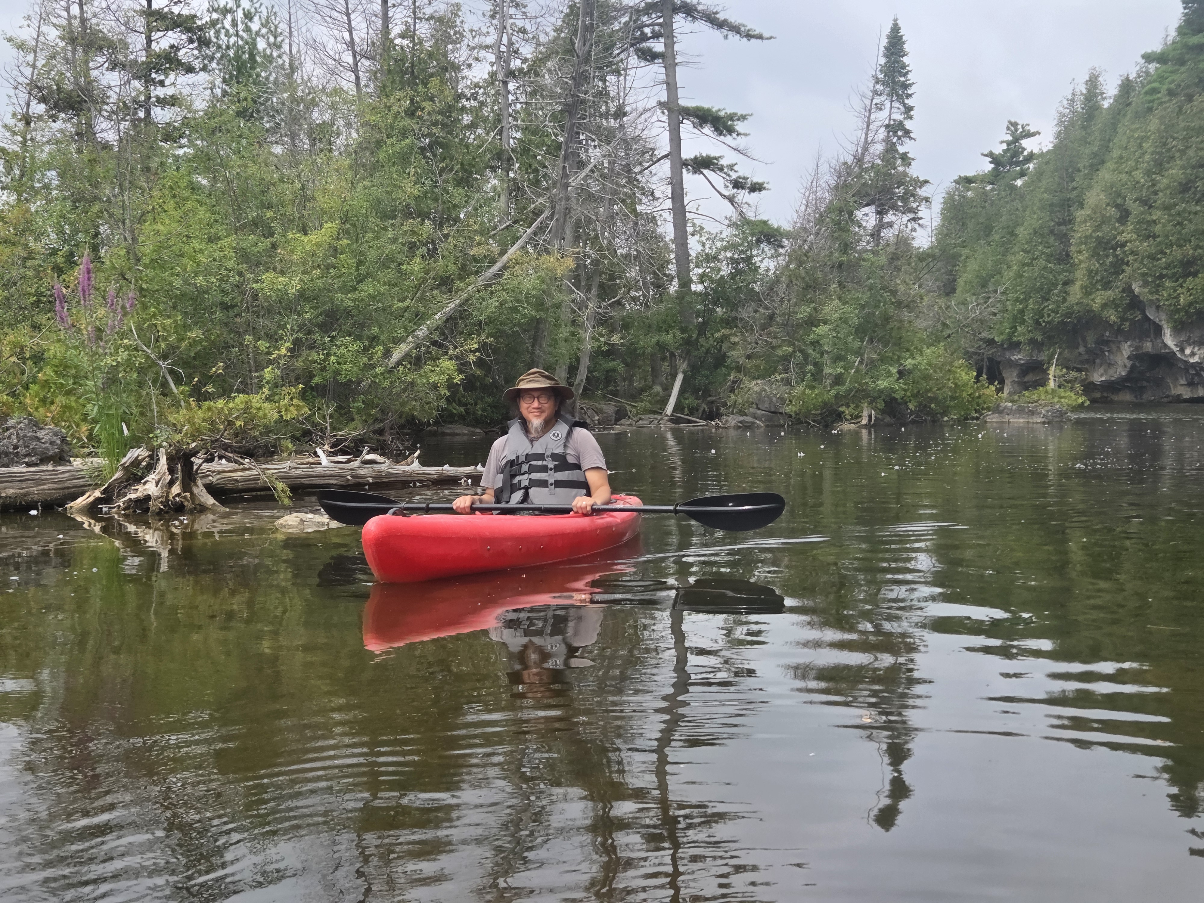 A person sitting in a red kayak on a calm waterway, surrounded by lush green trees and foliage, wearing a life jacket and a hat, with a paddle in hand.