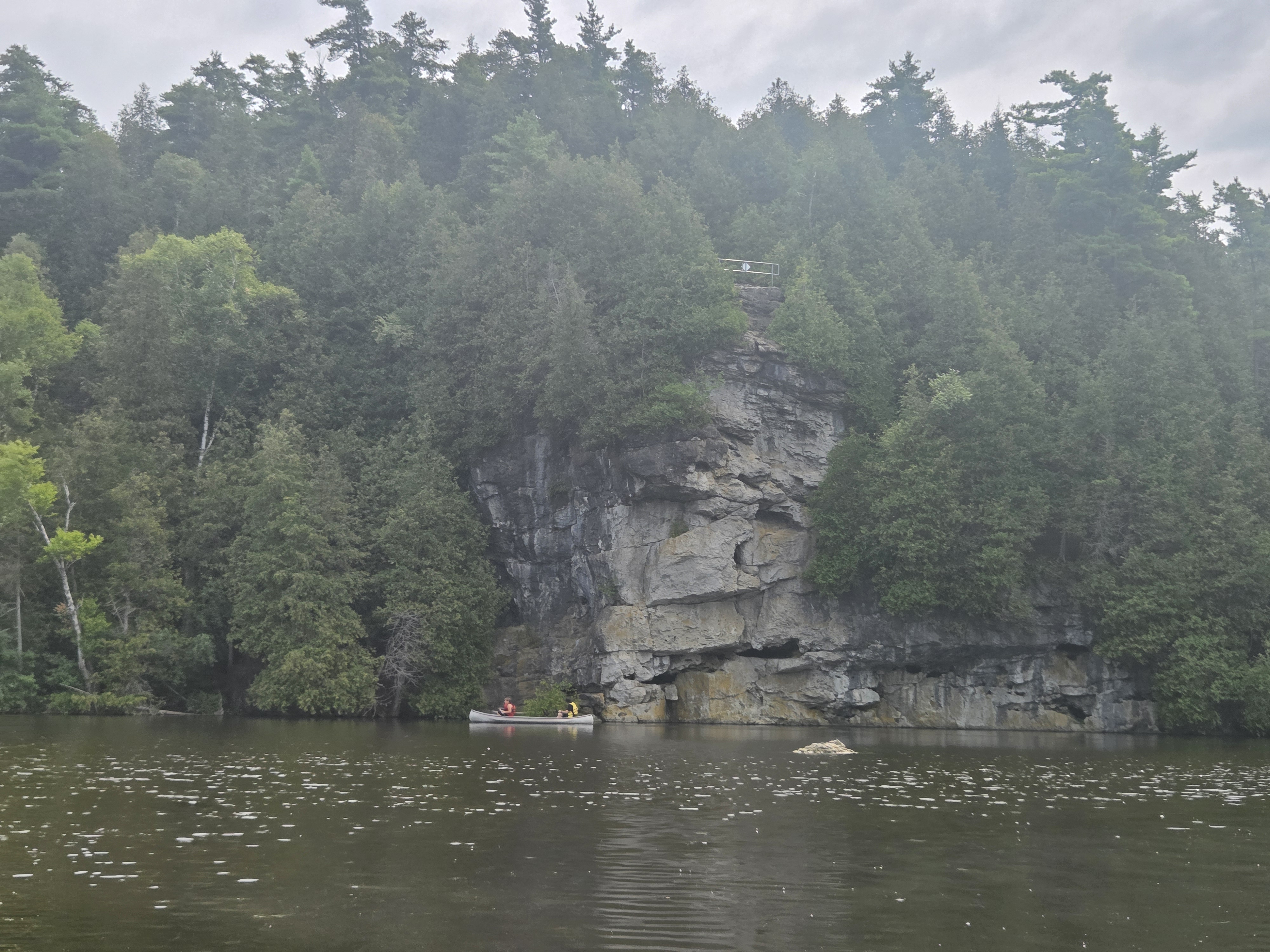 A serene lake scene featuring a canoe with two people paddling near a rocky cliff surrounded by dense green trees under a cloudy sky.