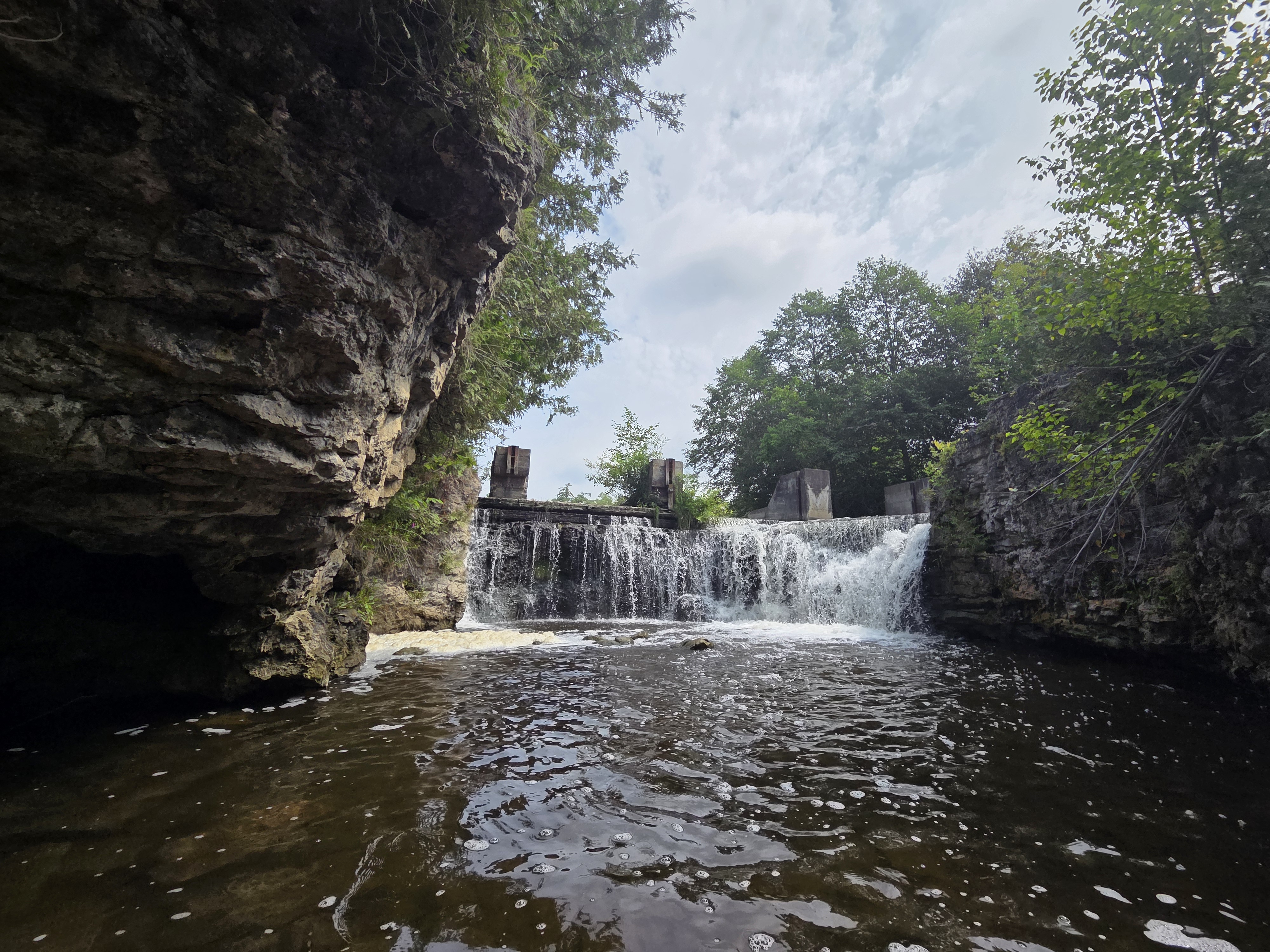 A scenic view of a waterfall flowing over a rocky ledge surrounded by trees, with a dam structure visible in the background.