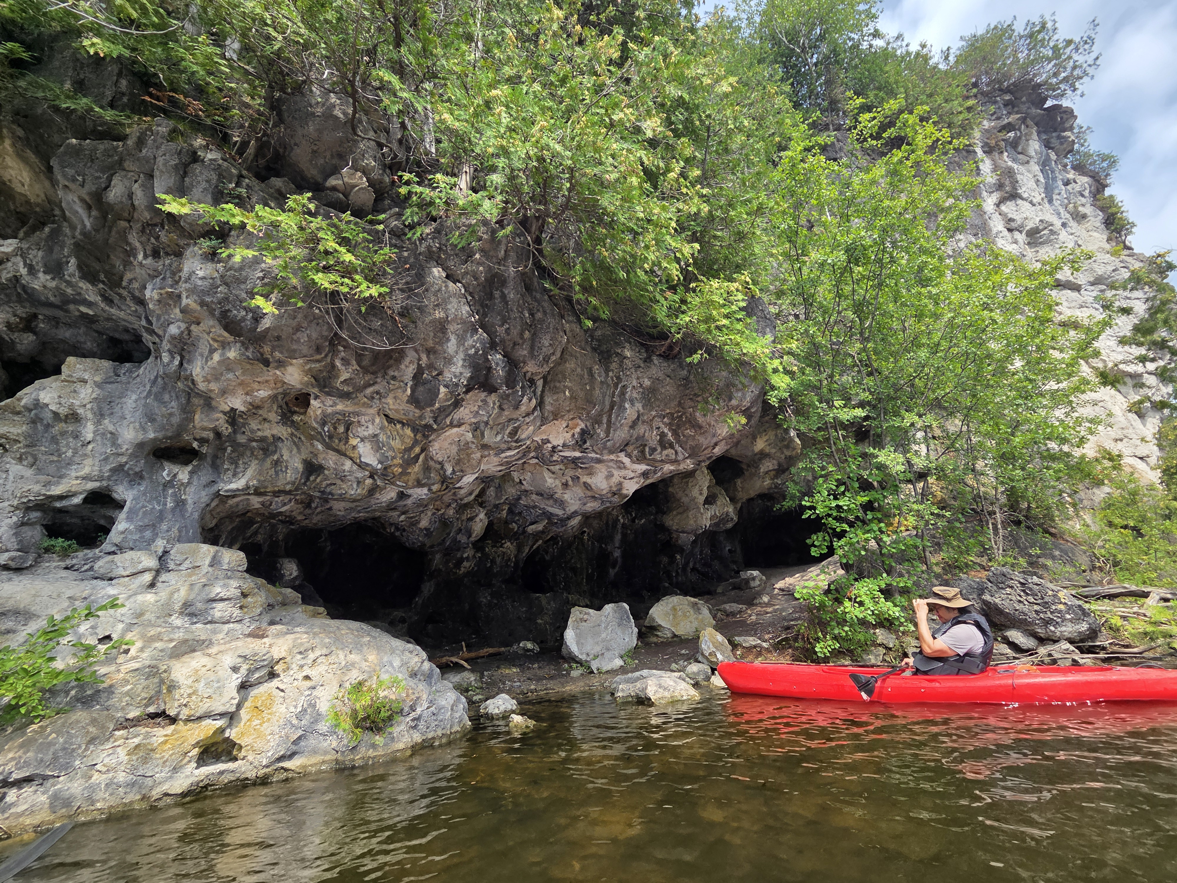 A person in a kayak is exploring a rocky shoreline with caves, surrounded by lush greenery and calm water.