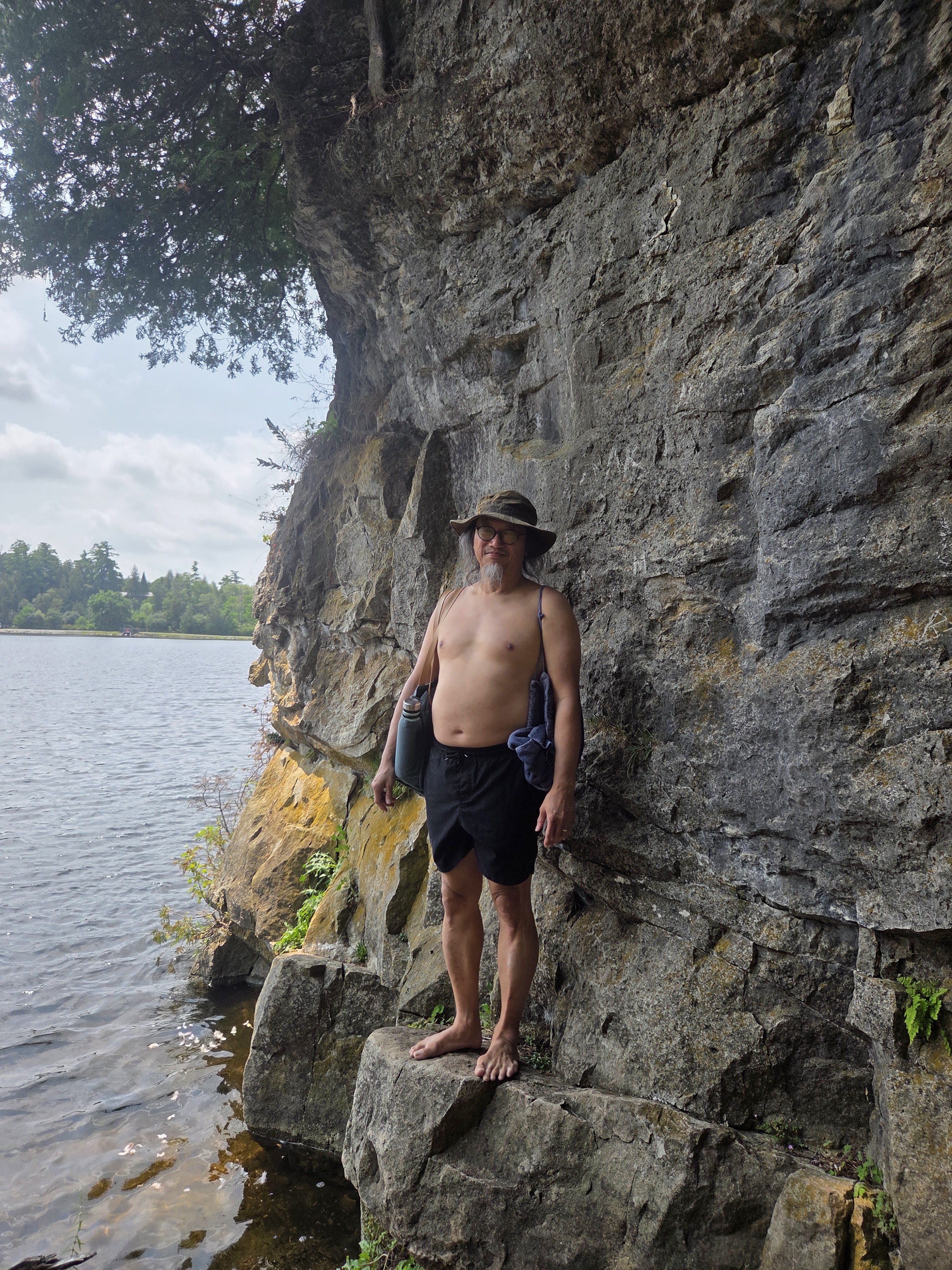 A man stands on a rock by the water, wearing shorts and a hat, with a towel and water bottle in his hands, against a rocky cliff and a tranquil lake background.