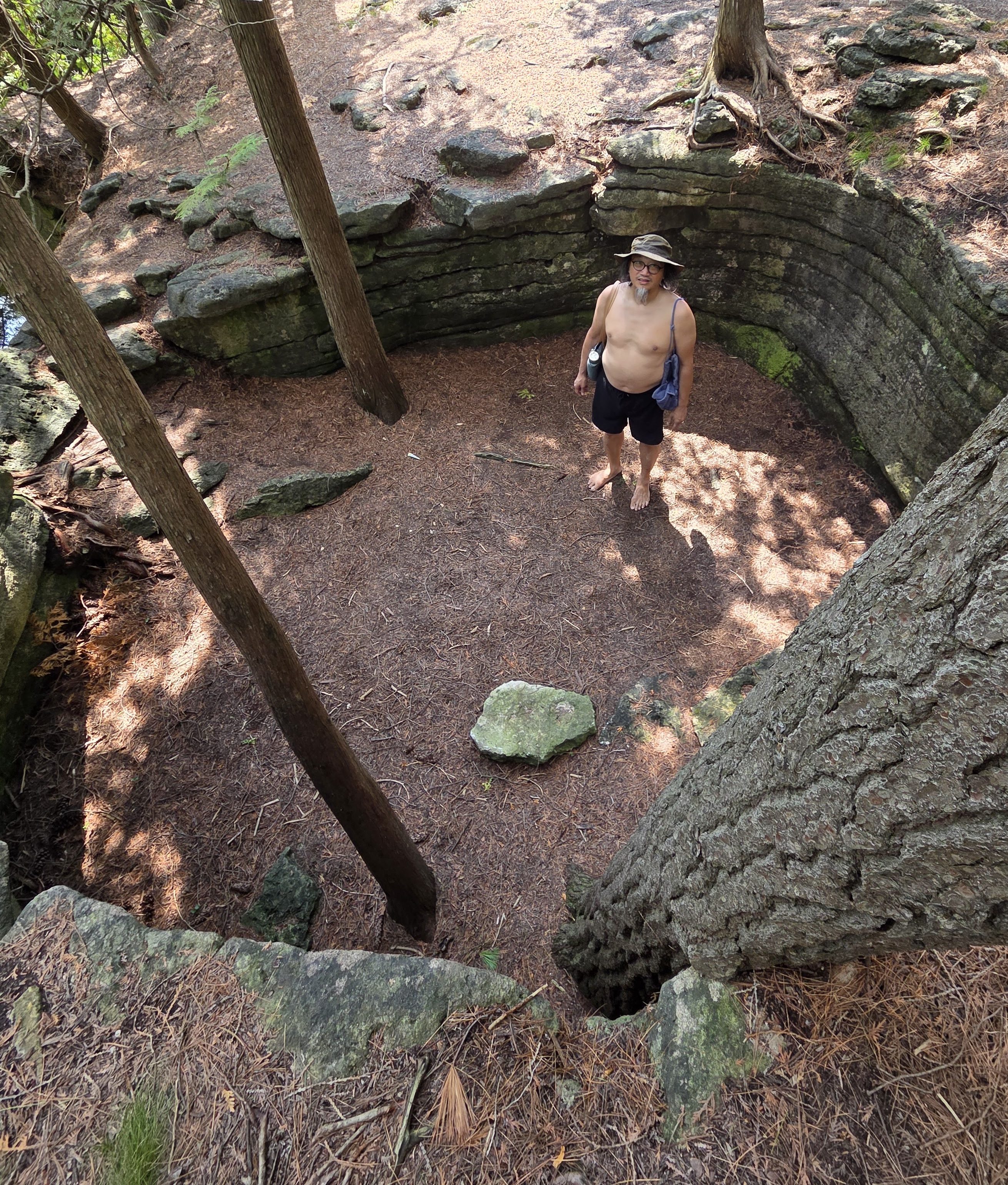 A shirtless man with a hat standing in a sunlit, circular stone pit surrounded by trees and rocks, with pine needles covering the ground.