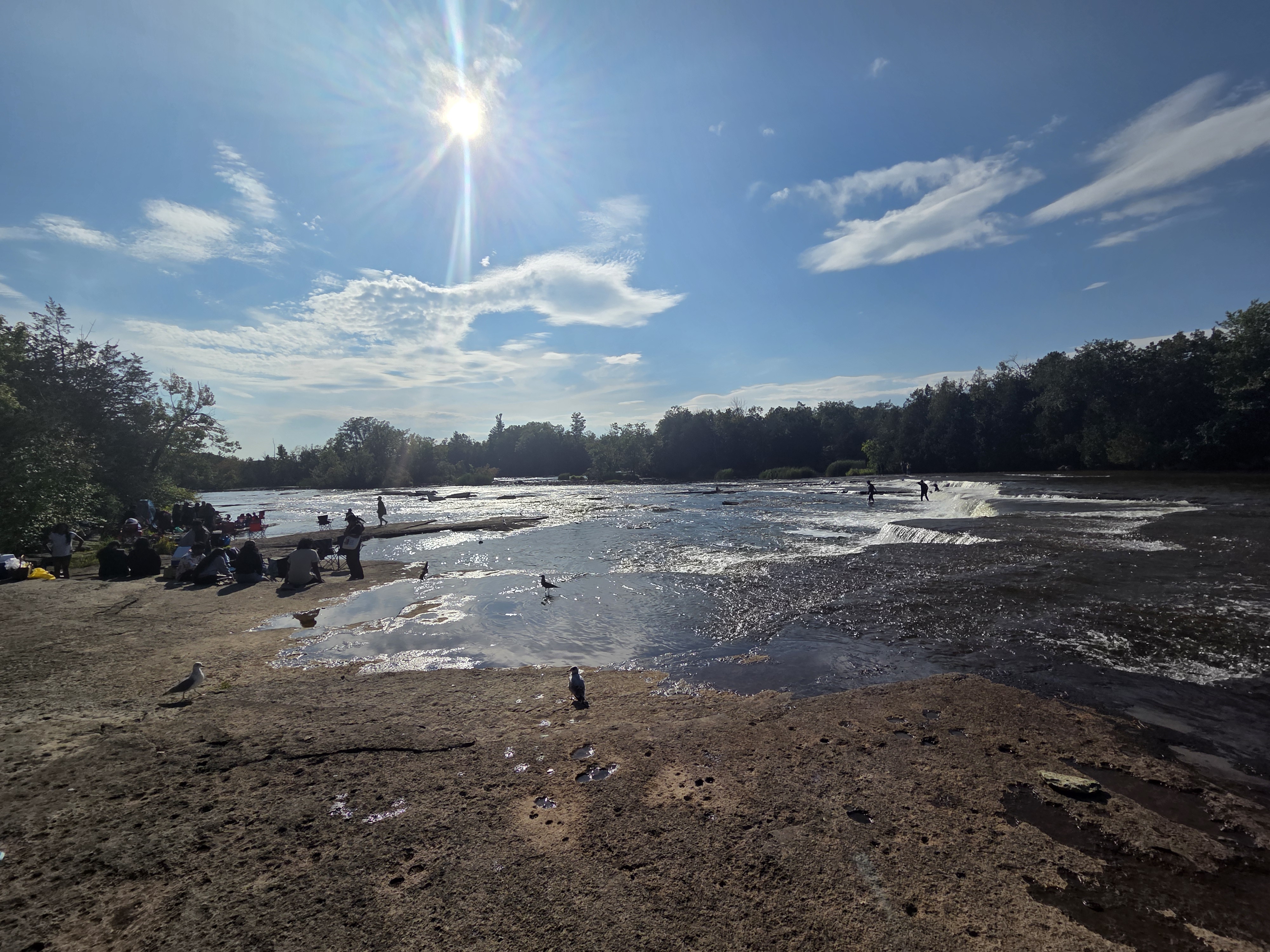 A sunny day at a riverbank with people gathered on the rocks, enjoying the water and scenery. The sun shines brightly in a clear blue sky, while a few individuals can be seen interacting near the water's edge.