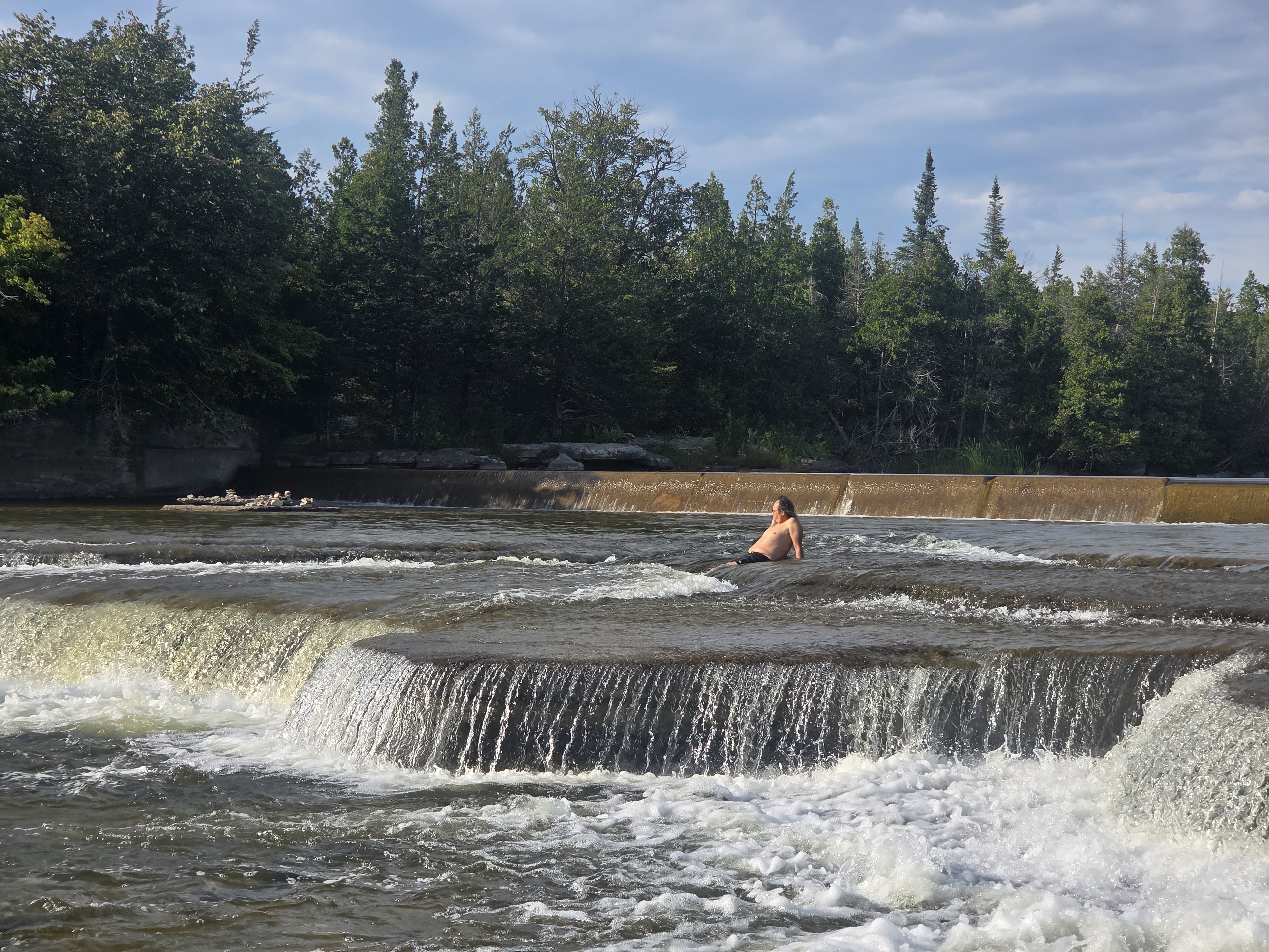 A person with long hair sitting on a rock in a river, surrounded by flowing water and greenery. Water cascades over small rock ledges in the background, with trees lining the riverbanks.
