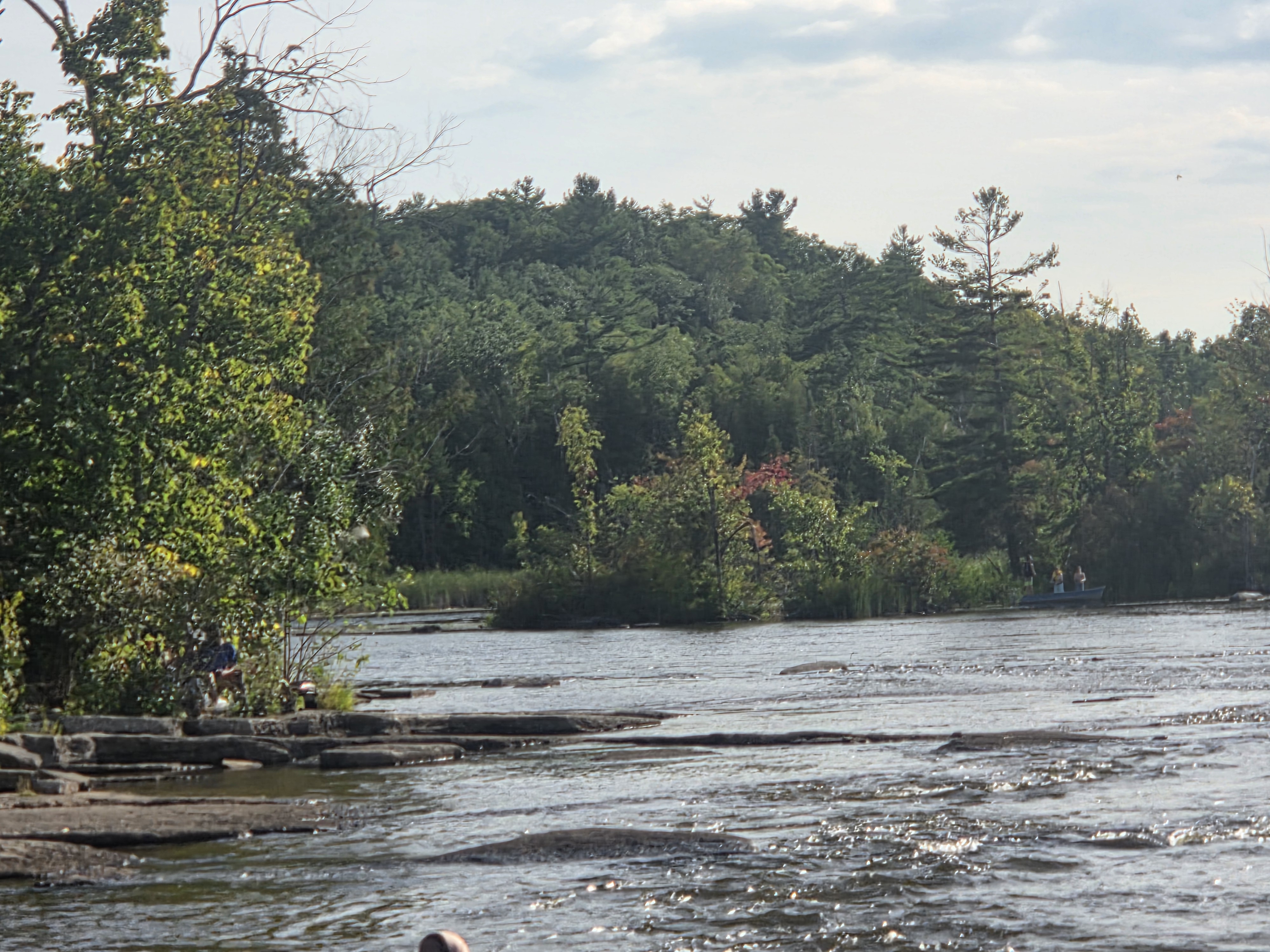 A tranquil river scene featuring rocky shores and lush green trees in the background, with a small boat carrying two people visible on the water.
