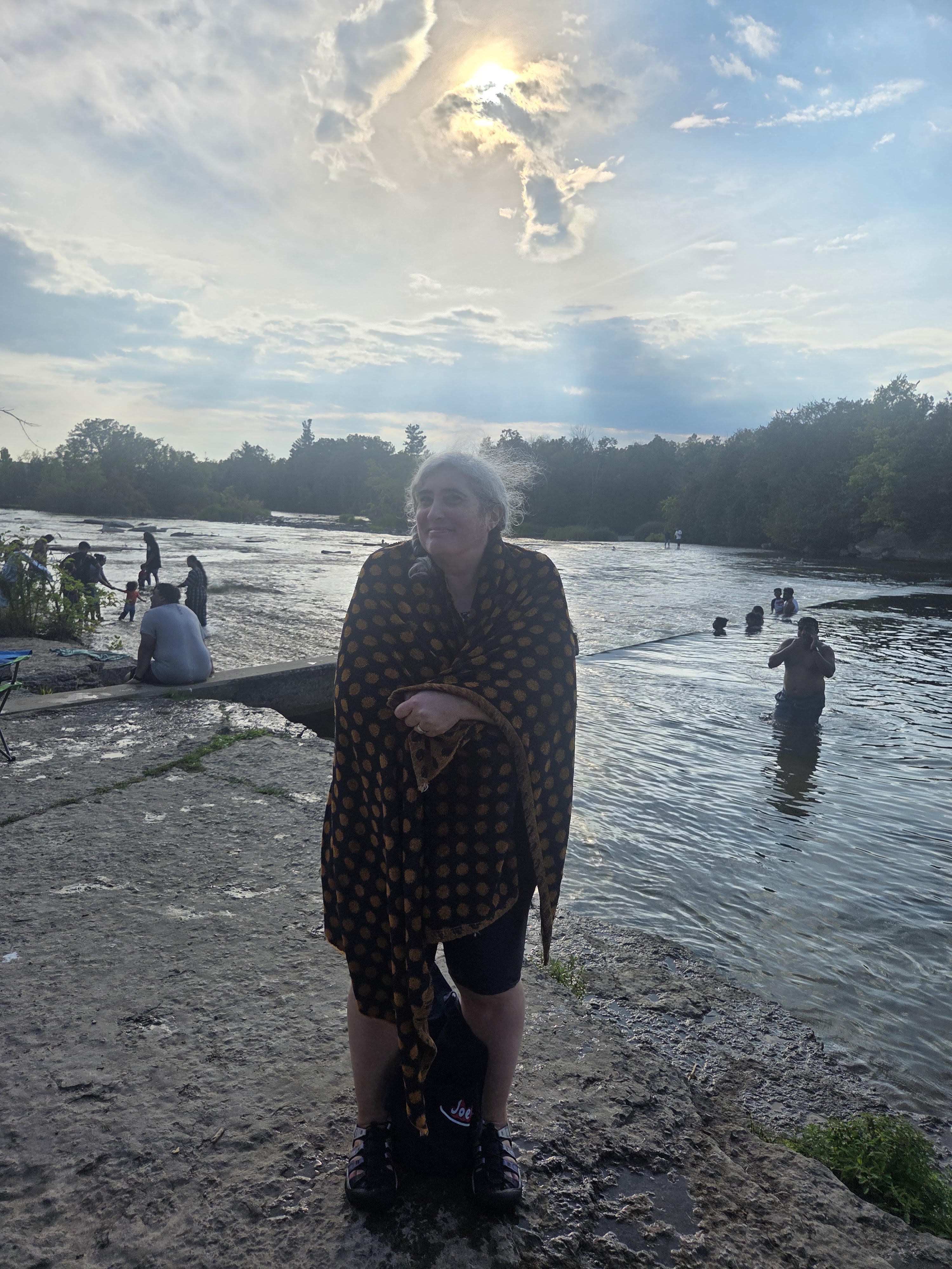 A woman standing on a rocky shore beside a river, wrapped in a patterned shawl, with cloudy skies and sun peeking through in the background. Other people are seen enjoying the water.
